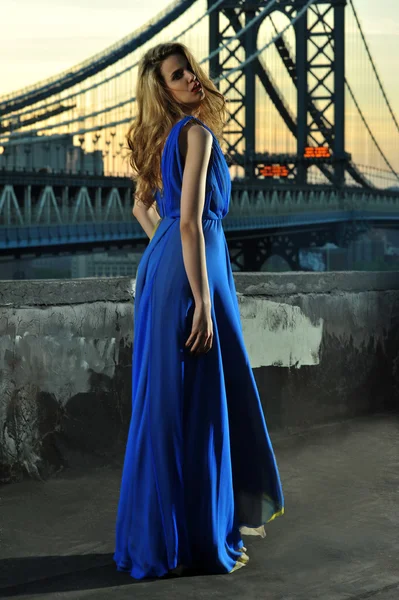 Fashion model posing sexy, wearing long blue evening dress on rooftop location with metal bridge construction on background