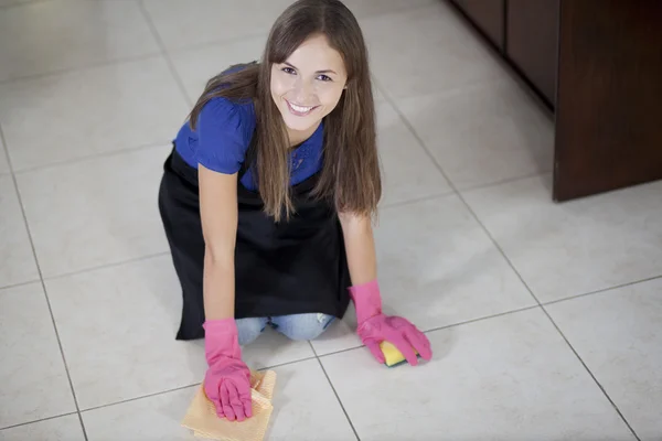 Gorgeous smiling woman in pink gloves and black apron cleaning the floor while kneeling at home