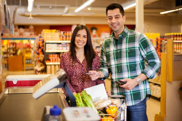 Cute young couple standing with a trolley with products in the store at checkout and man paying with credit card