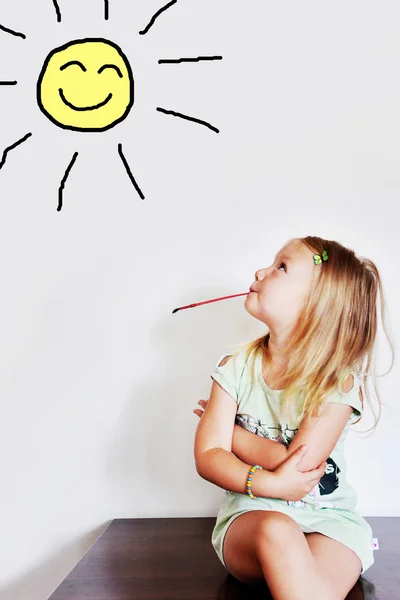 Girl sitting on a table with a brush in his mouth and looks at the sun painted on the wall