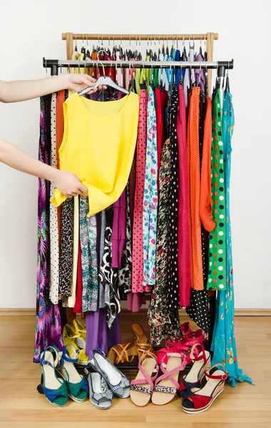 Woman hand picking up a yellow blouse to wear.  Summer dresses and sandals in a wardrobe.