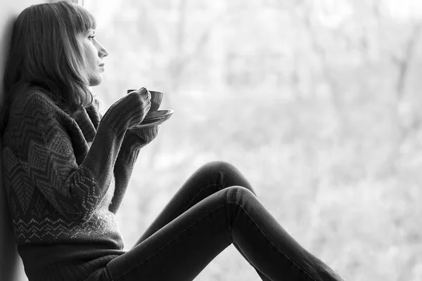 Beautiful girl dreaming with cup of coffee or tea near window. Black and White image