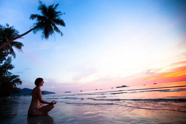 Beautiful woman doing lotus yoga pose on the beach near the ocean at sunset in Thailand