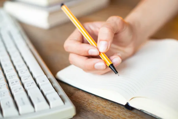 Hands writes in a notebook, keyboard, a stack of books in background