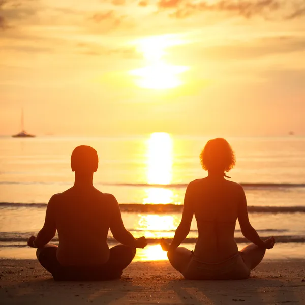 Yoga practice: a young couple sitting on the beach of the sea in the lotus position at sunset.