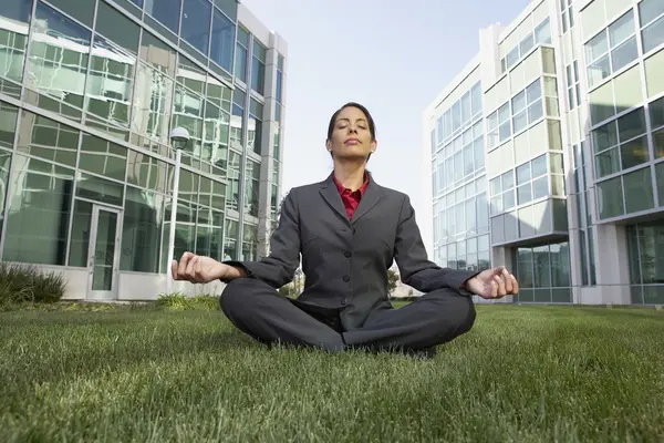 Hispanic businesswoman meditating in the grass in front of office buildings