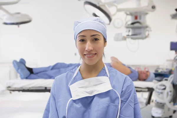 Hispanic female doctor smiling in operating room with male doctor laying on table in background