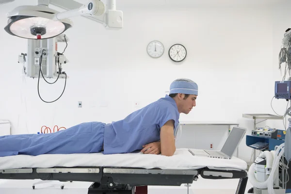 Male doctor laying on table in operating room using laptop
