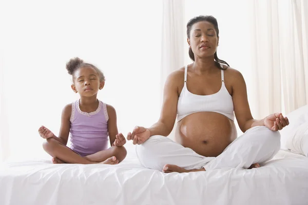 Pregnant African mother with young daughter sitting on the bed meditating