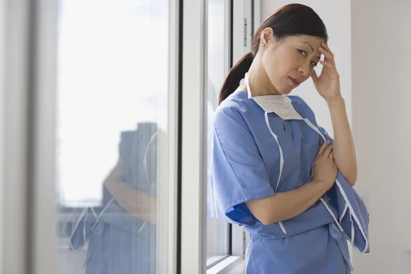 Asian female doctor leaning against window with hand on forehead