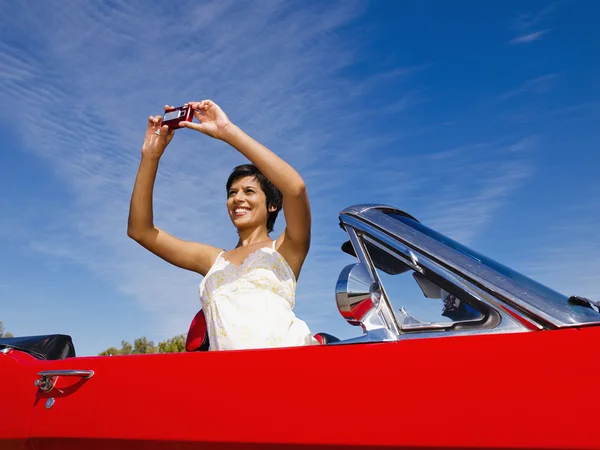 Mixed race woman taking photograph from red convertible