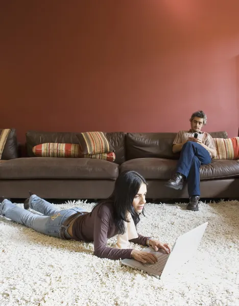 Woman laying on floor using laptop and man sitting on sofa using cell phone
