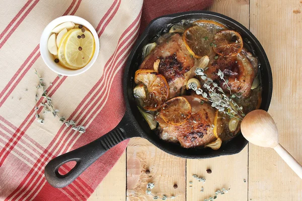 Baked chicken thighs with lemon slices, dried thyme, garlic cloves, black peppercorns and white wine sauce in a pan on a wooden table