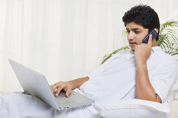 Young man using cell phone while working on laptop