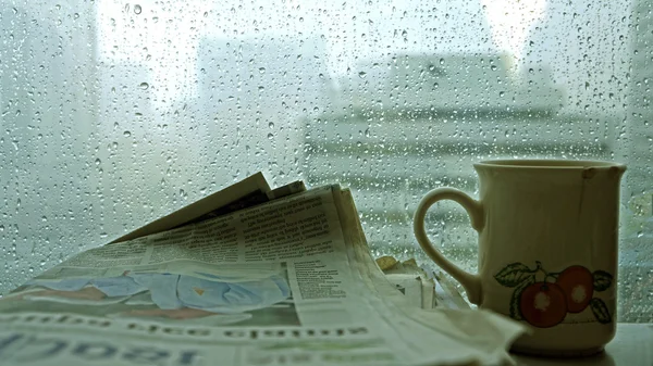 Cup of coffee with newspaper in dark room with rain outside