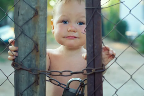 Upset baby boy looking out of locked wire fencing. outdoors