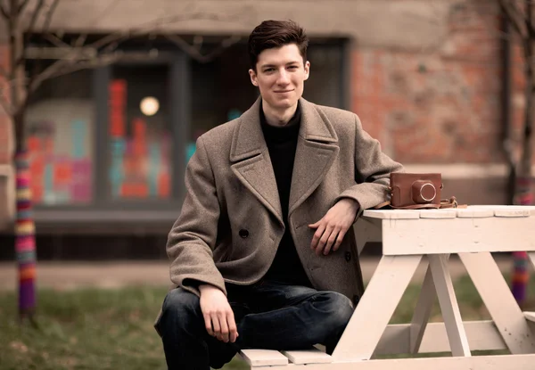 The young man model in a coat sits at a white table with the vintage camera outdoors