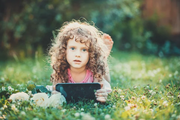 Little girl sitting on grass and hold white flowers and tablet p