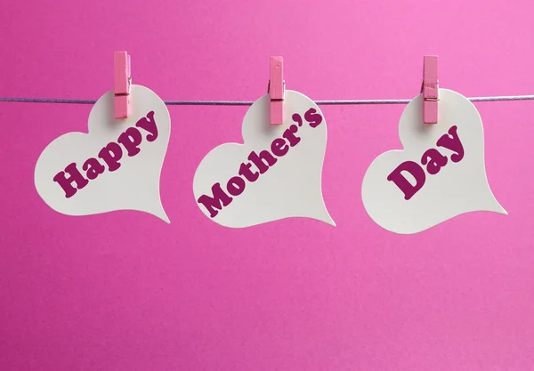 Happy Mothers Day message written across white heart shape gift tags hanging from pegs on a line against a pink background.