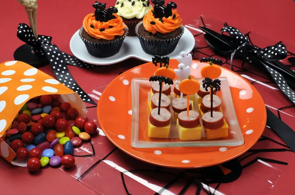 Happy Halloween party table with skeleton glass, cupcakes, candy lollies and party food with orange and black pumpkin, cat, bat and ghost decorations.