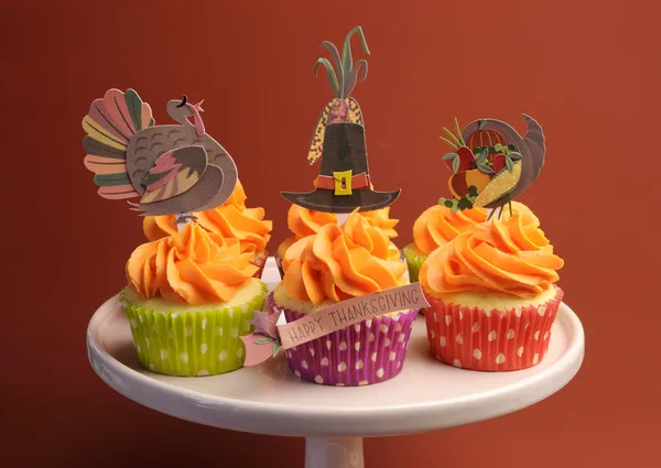 Happy Thanksgiving decorated cupcakes with turkey, pilgrim hat and corn toppers on cake stand against a brown background.