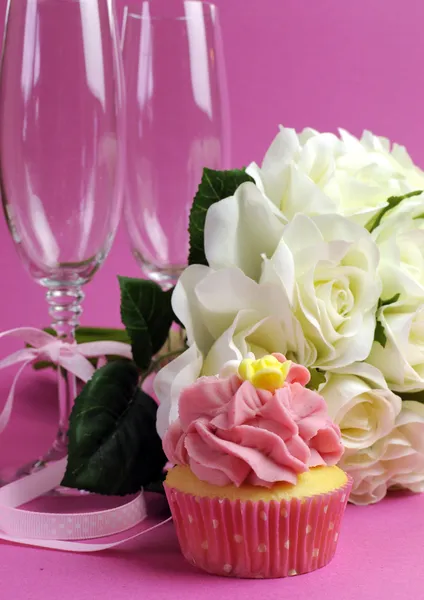 Wedding bridal bouquet of white roses on pink background with pink cupcake and pair of two champagne flute glasses. Vertical.