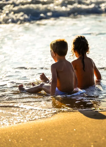 Two young children sitting in the waves of the ocean under a beautiful sunset