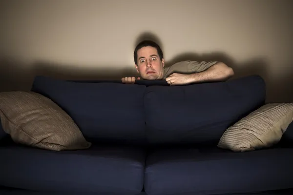 Frightened man hides behind couch watching TV