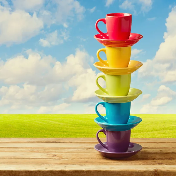 Rainbow color cups on wooden table over beautiful sky and meadow.