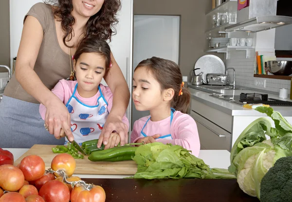 Mum and twin daughters learning to chop vegetables together in the kitchen, using a chopping board and surrounded by fruit and vegetables.