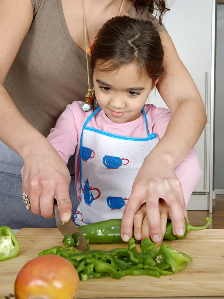 Mother teaching daughter to chop vegetables together in the kitchen using a chopping board and smiling.