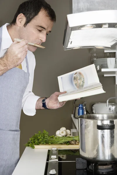 Businessman tasting his food while using a recepe book to cook at home.