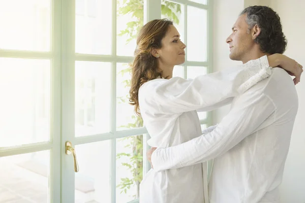 Portrait of a mature couple hugging at home, standing by large garden dooors.