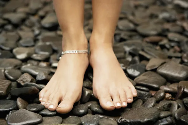 Woman\'s feet wearing an anklelet and standing on black stones.