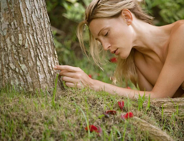 Beautiful blonde woman laying naked on green grass and tree roots.