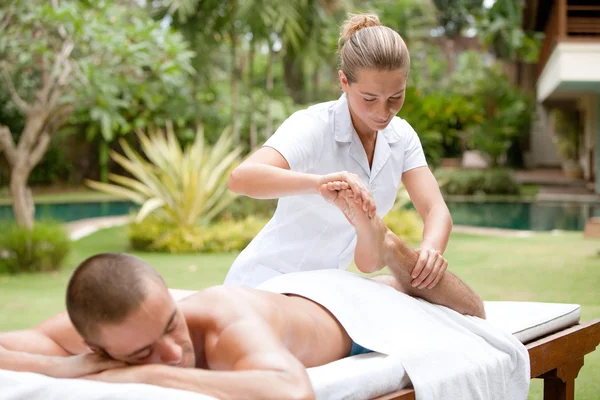 Young masseuse massaging and stretching the body of an attractive man in a tropical hotel garden