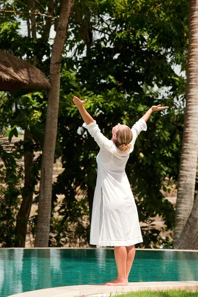 Young woman\'s figure with arms outstretched while standing at the edge of a swimming pool in a tropical nature location.