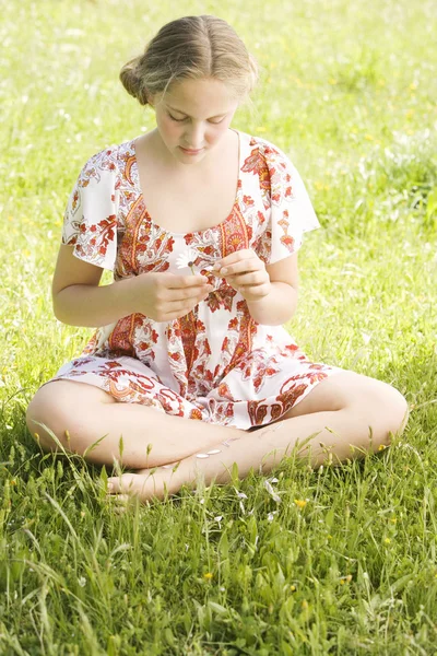 Girl pulling petals off a daisy flower while sitting on a green field, playing love me, love me not.