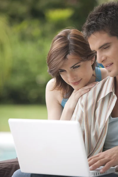 Man and woman looking at laptop screen, sitting by swimming pool in garden.