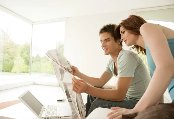 Young couple sharing a newspaper at home while sitting on the sofa.