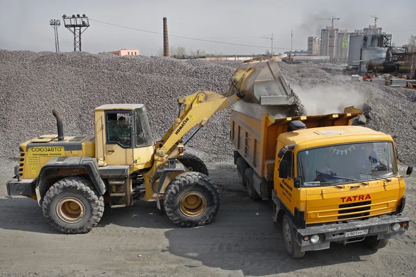 Yellow  excavator loads gravel into orange dumper truck tipper.