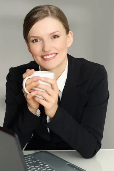 Happy woman sitting in armchair with laptop computer, holding coffee mug, looking at camera