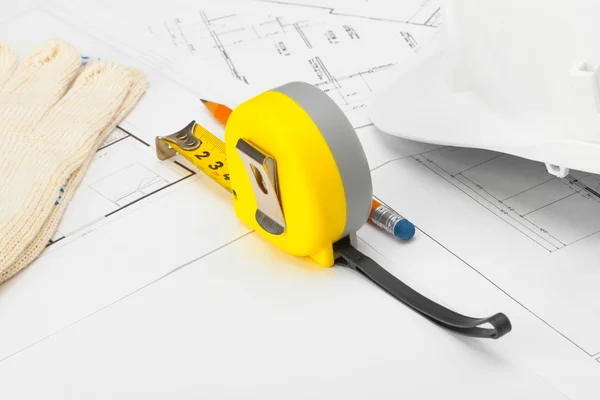Yellow measure tape with gloves, construction helmet and pencil above blueprint