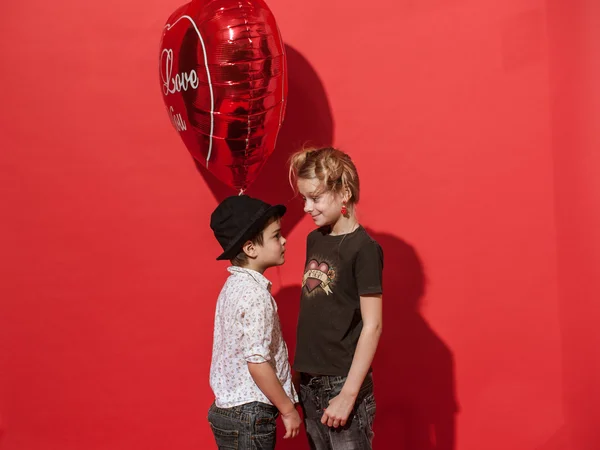 Girl and boy with red balloon on the red background, they are smiling and trying to kiss each other.