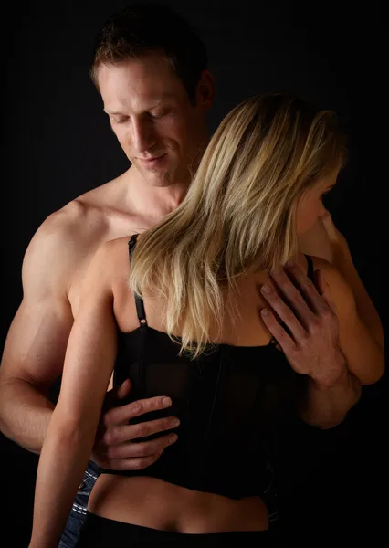 Young and fit caucasian adult couple in an embrace. Semi-nude and topless against a dark background with the woman wearing a sexy red and black lace corset..