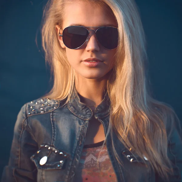 Outdoor summer closeup portrait of young stylish fashion glamorous woman or girl in sunny day on street jeans jacket and sunglasses standing near blue wall