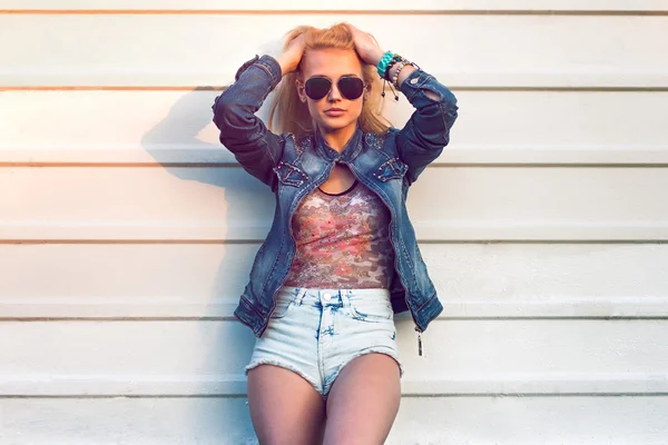 Outdoor summer closeup portrait of young stylish fashion glamorous woman or girl posed in  sunny day on street jeans jacket and sunglasses standing near white wall