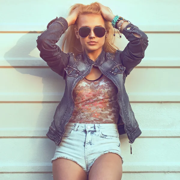 Outdoor summer closeup portrait of young stylish fashion glamorous woman or girl posed in  sunny day on street jeans jacket and sunglasses standing near white wall