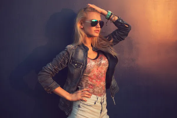 Outdoor summer closeup portrait of young stylish fashion glamorous woman or girl in  sunny day on street jeans jacket and sunglasses standing near blue wall