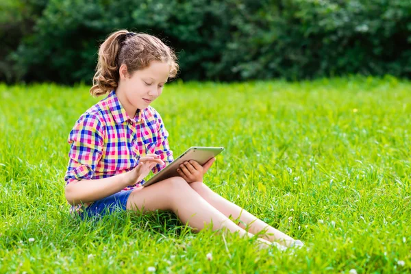 Outdoor portrait of a pretty teenager girl in casual clothes sitting on the grass with digital tablet on her knees, reading and surfing